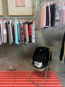Clothing rack with various garments on a concrete floor, with a black bag on a chair in the foreground.