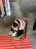 Pink backpack with a shoe inside on a chair, with clothes hanging in the background.